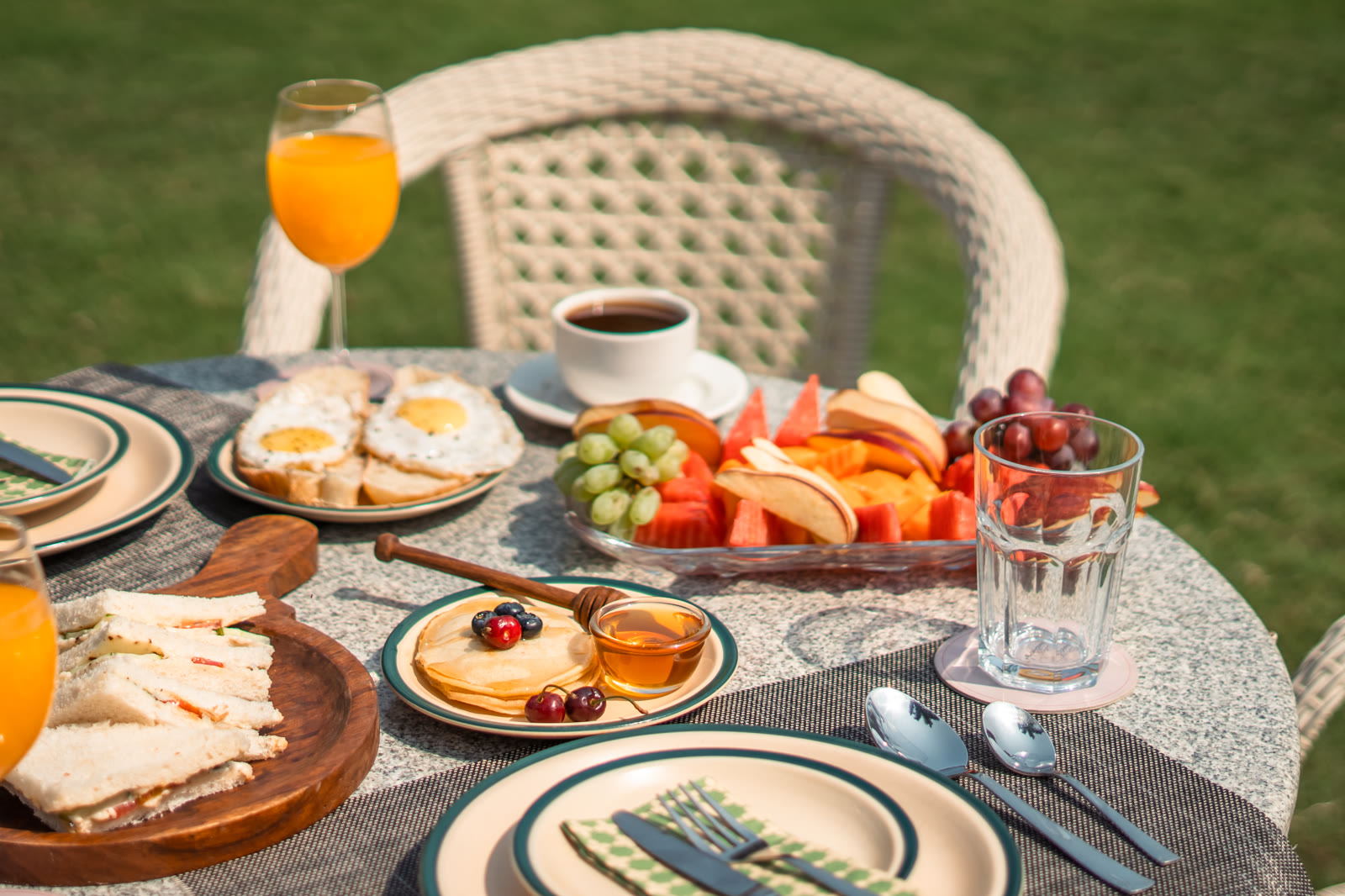 Breakfast on a table on the lawn: fried eggs on toast, pancakes with honey and berries, cut fruit, sandwiches, orange juice and coffee